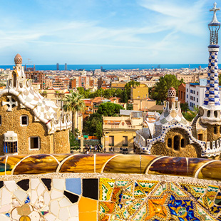 Park Güell in Barcelona met kleurrijke mozaïekbank, sierlijke vormen en uitzicht over de stad met palmbomen en gebouwen op de achtergrond.
